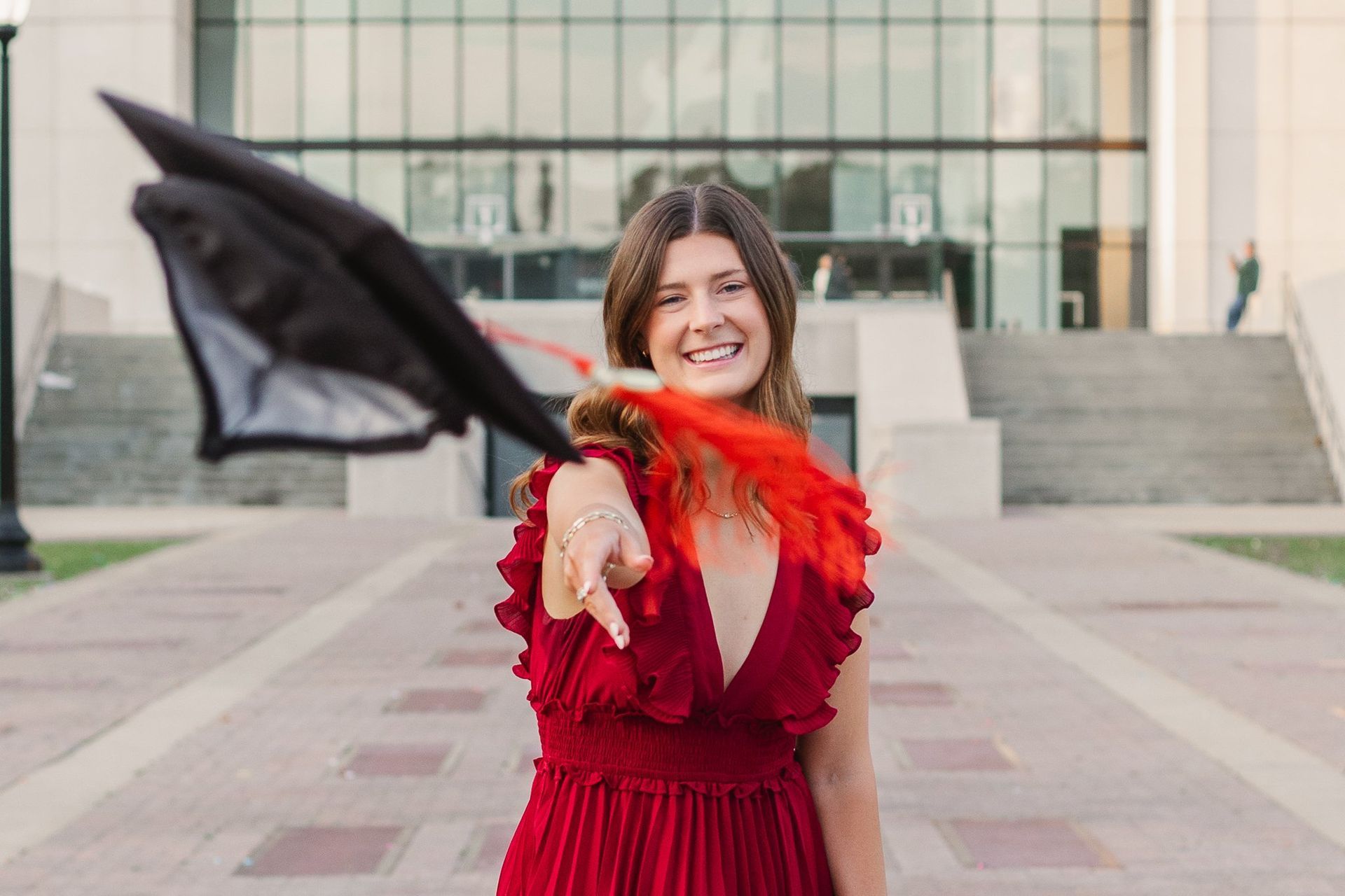 A woman in a red dress is holding a graduation cap and pointing at the camera.