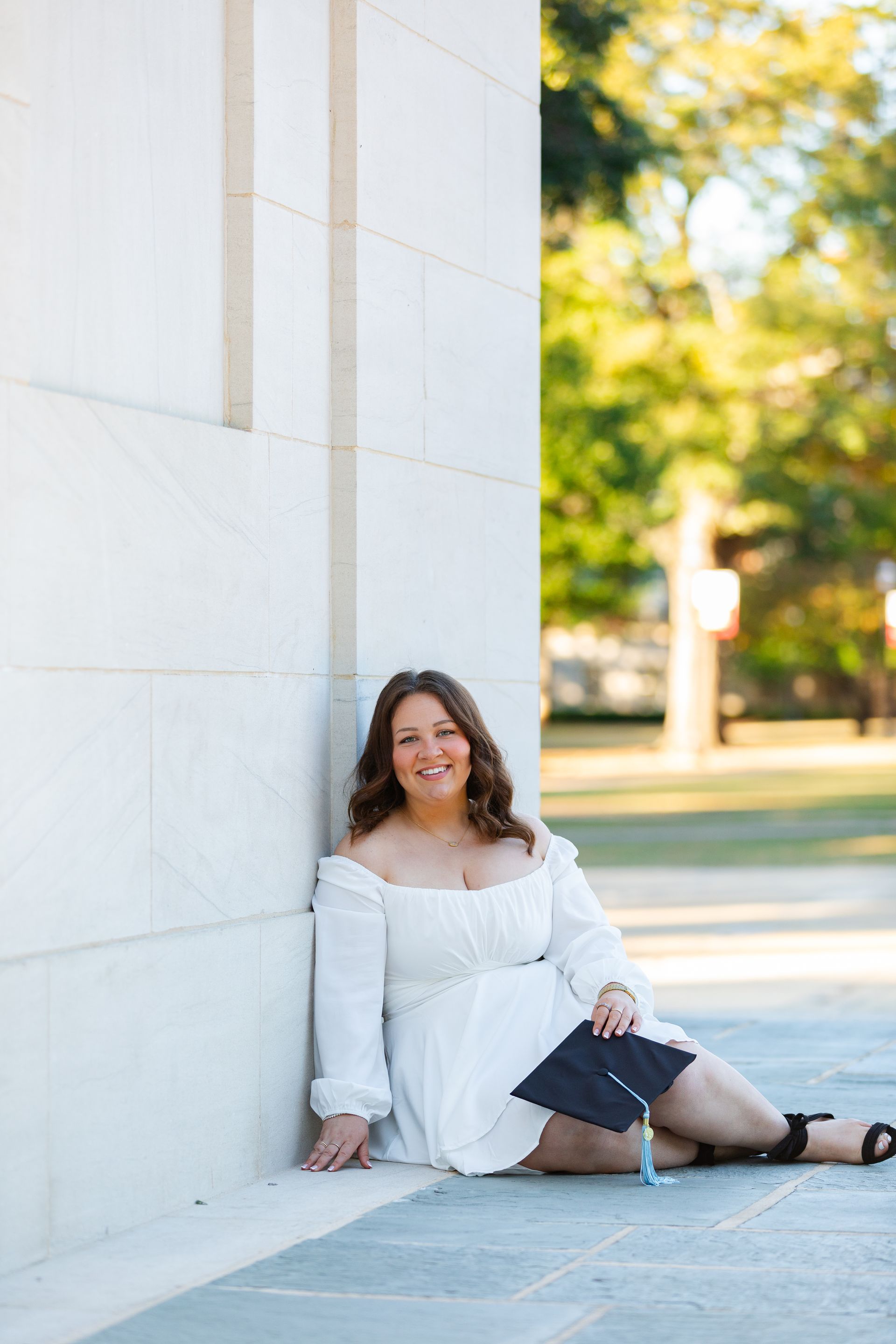 A woman in a white dress is sitting on the ground leaning against a wall.
