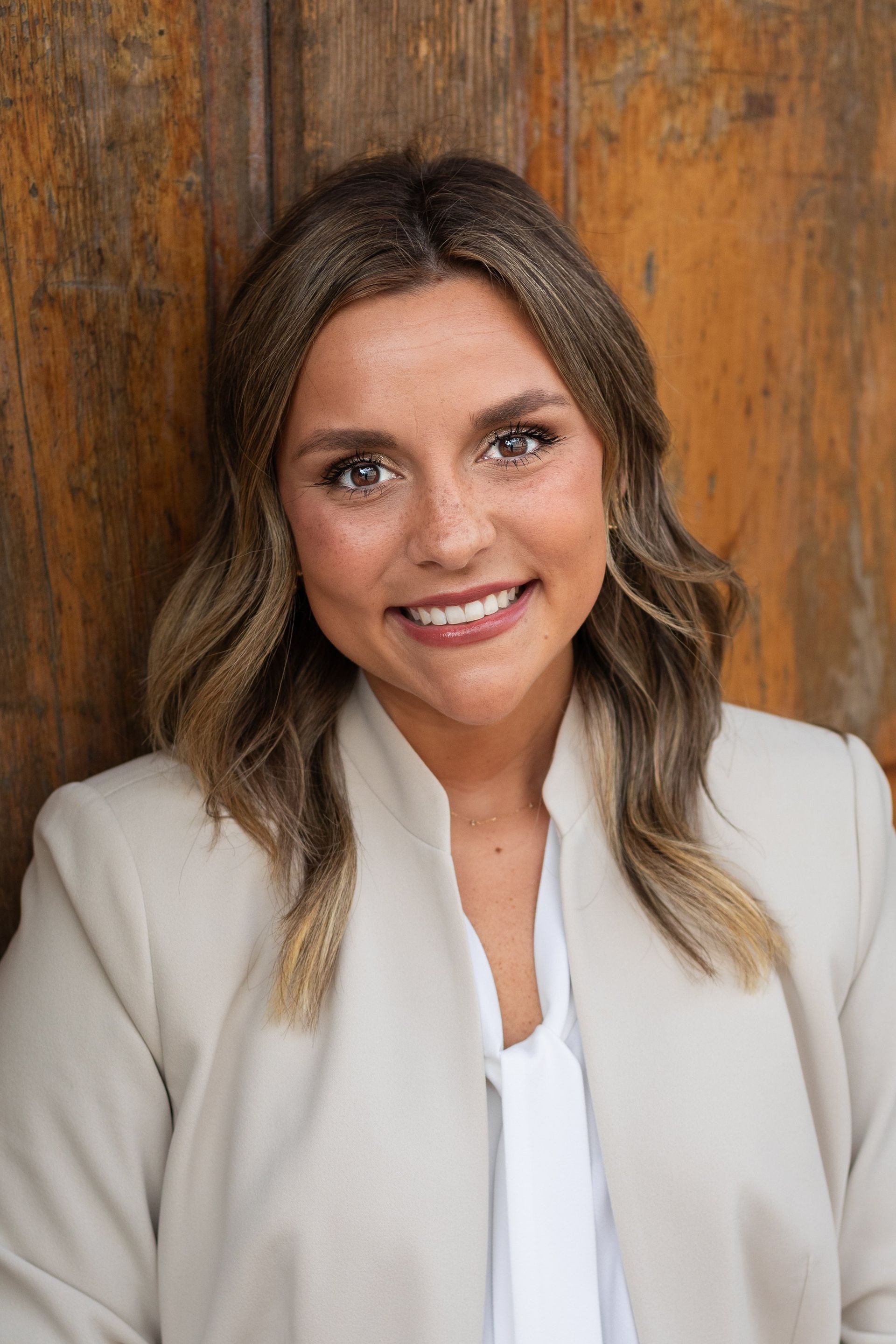 A woman in a white jacket and white shirt is smiling in front of a wooden wall.