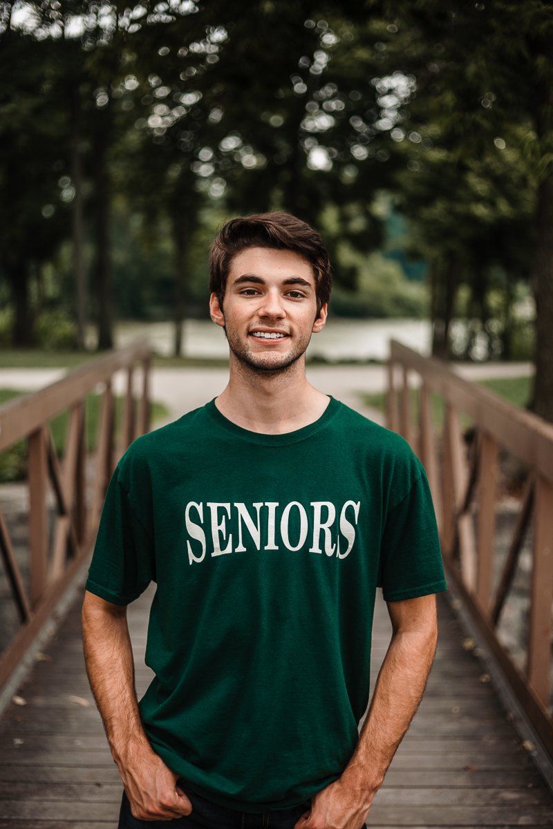A young man wearing a green seniors t-shirt is standing on a bridge.
