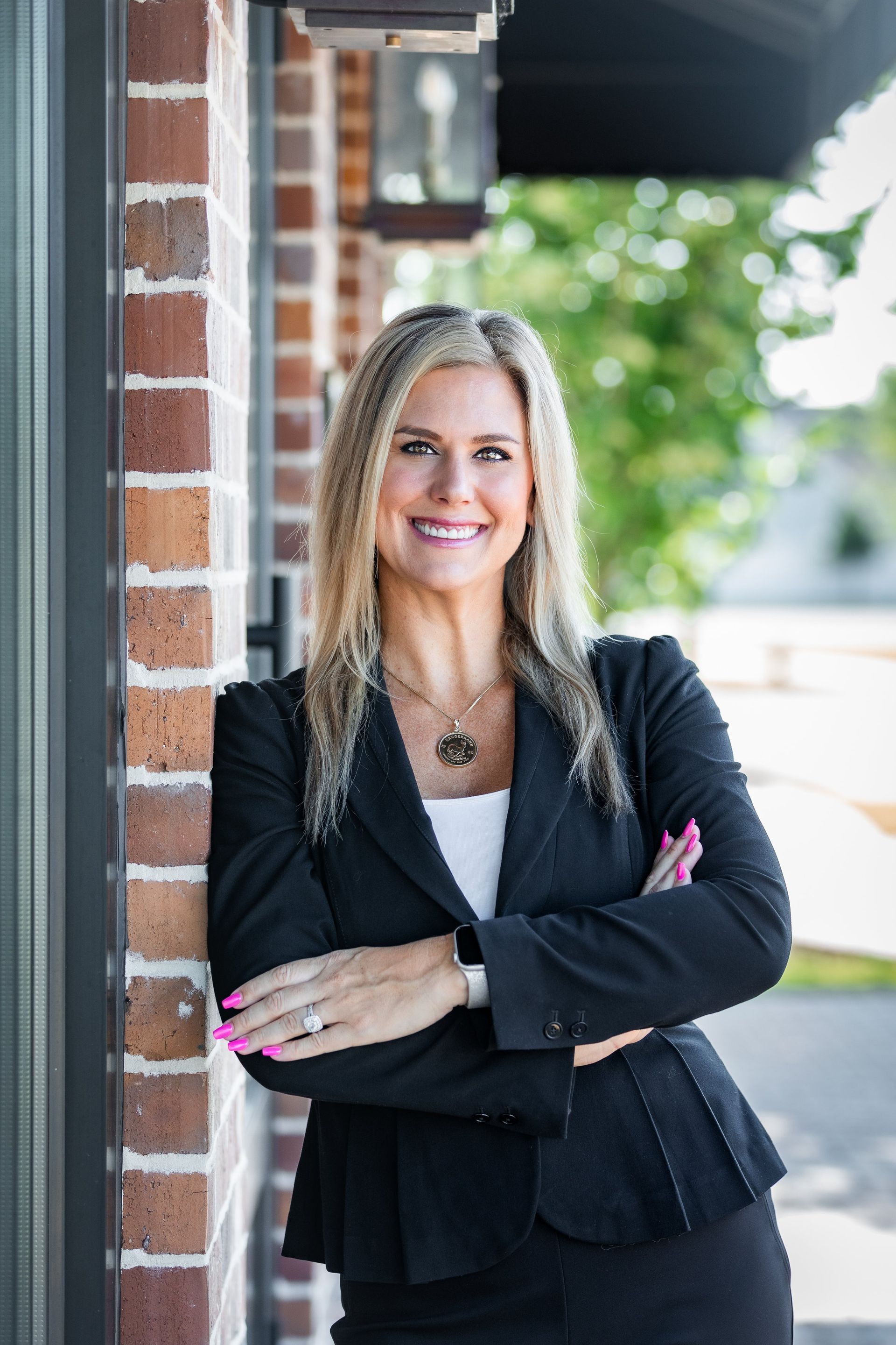 A woman in a black suit is standing in front of a brick building with her arms crossed.