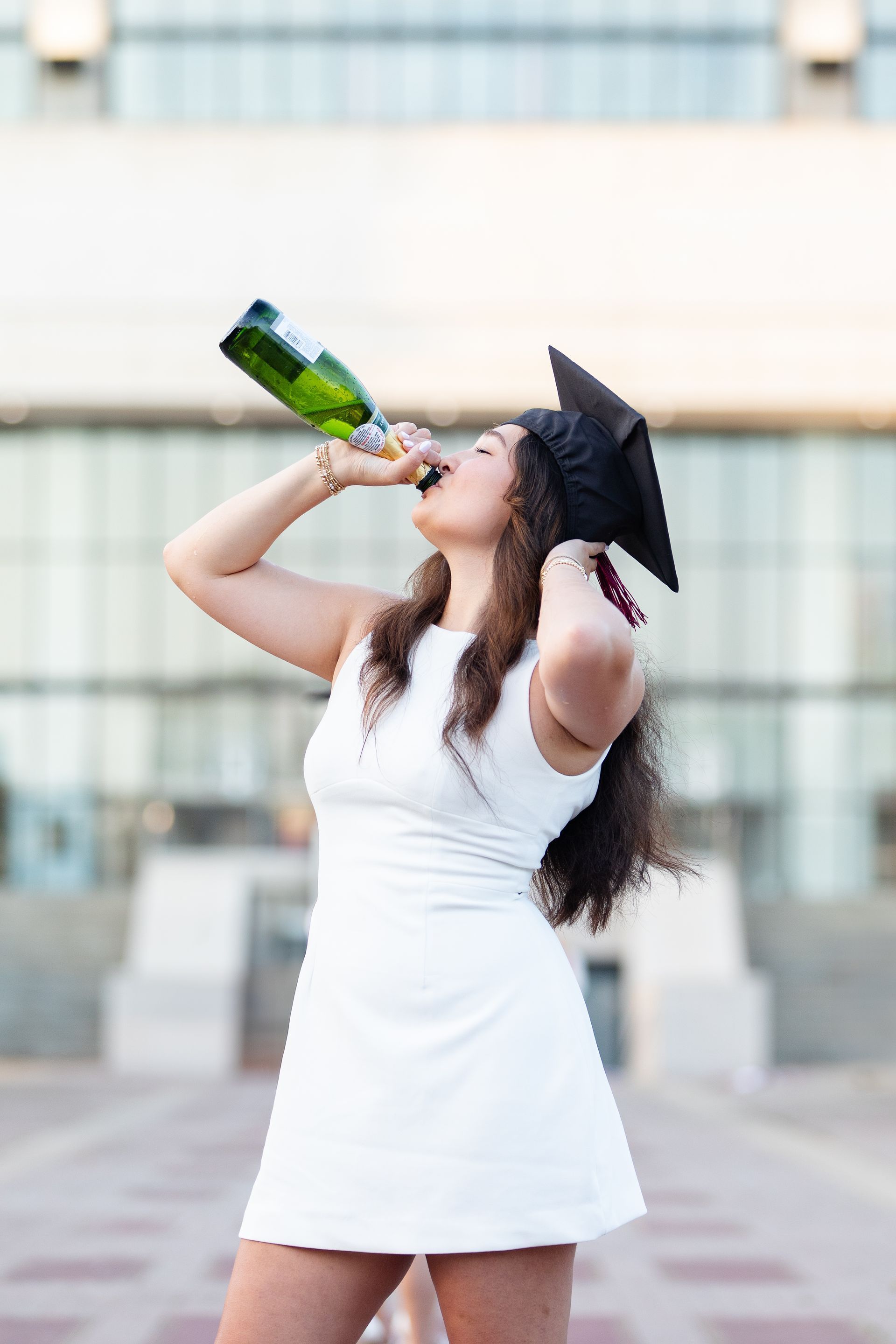 A woman in a graduation cap and gown is drinking from a bottle of champagne.