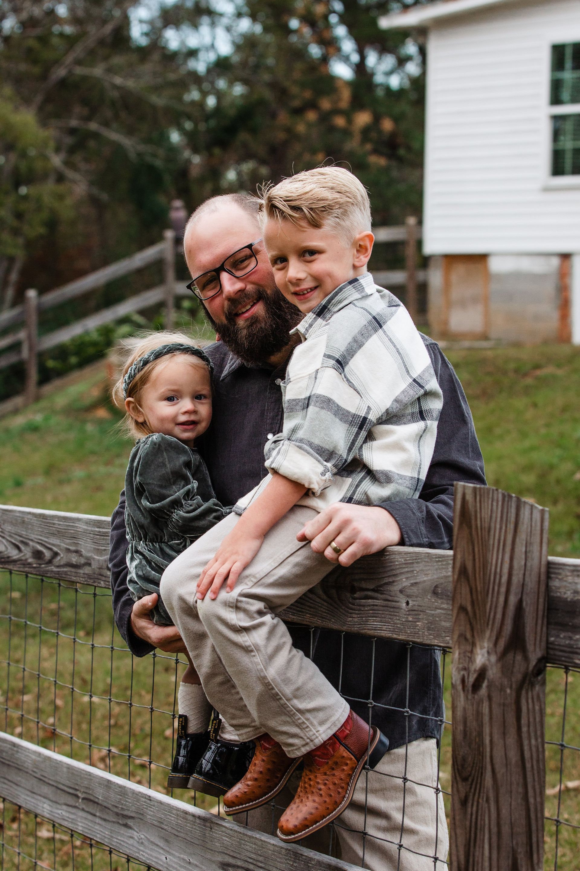 A man is holding two children while sitting on a wooden fence.