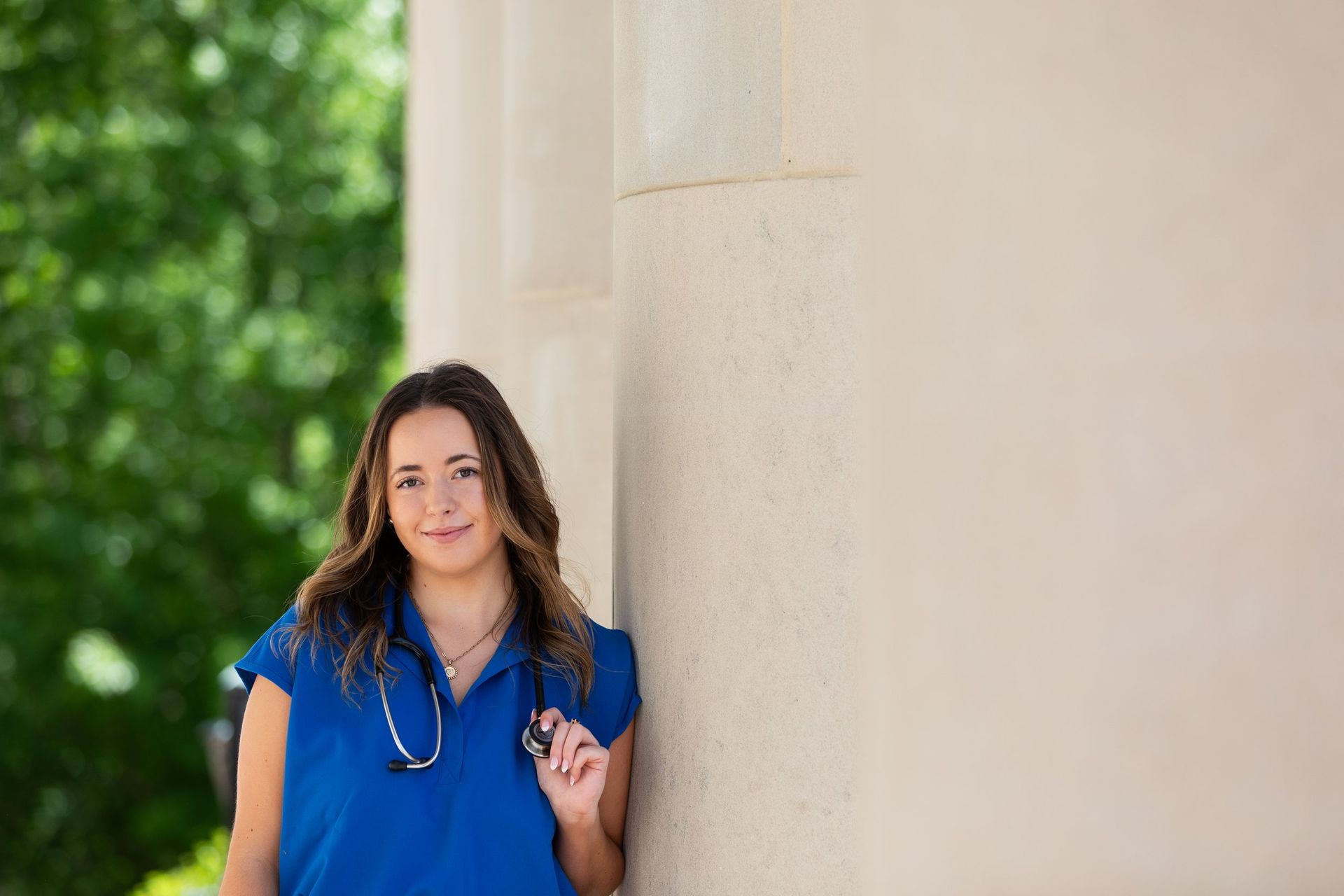 A woman in a blue shirt and stethoscope is leaning against a wall.