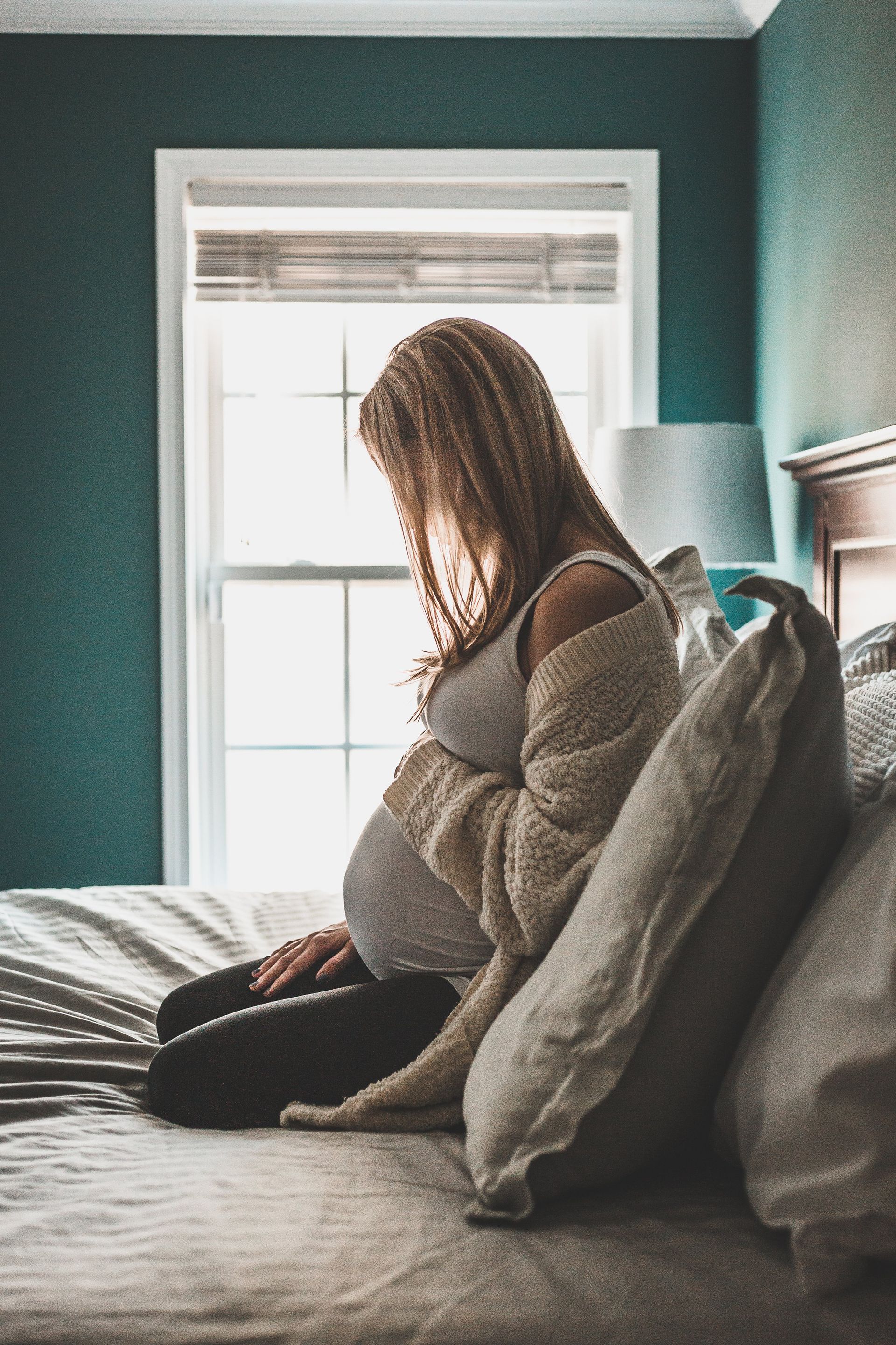 A pregnant woman is sitting on a bed in front of a window.