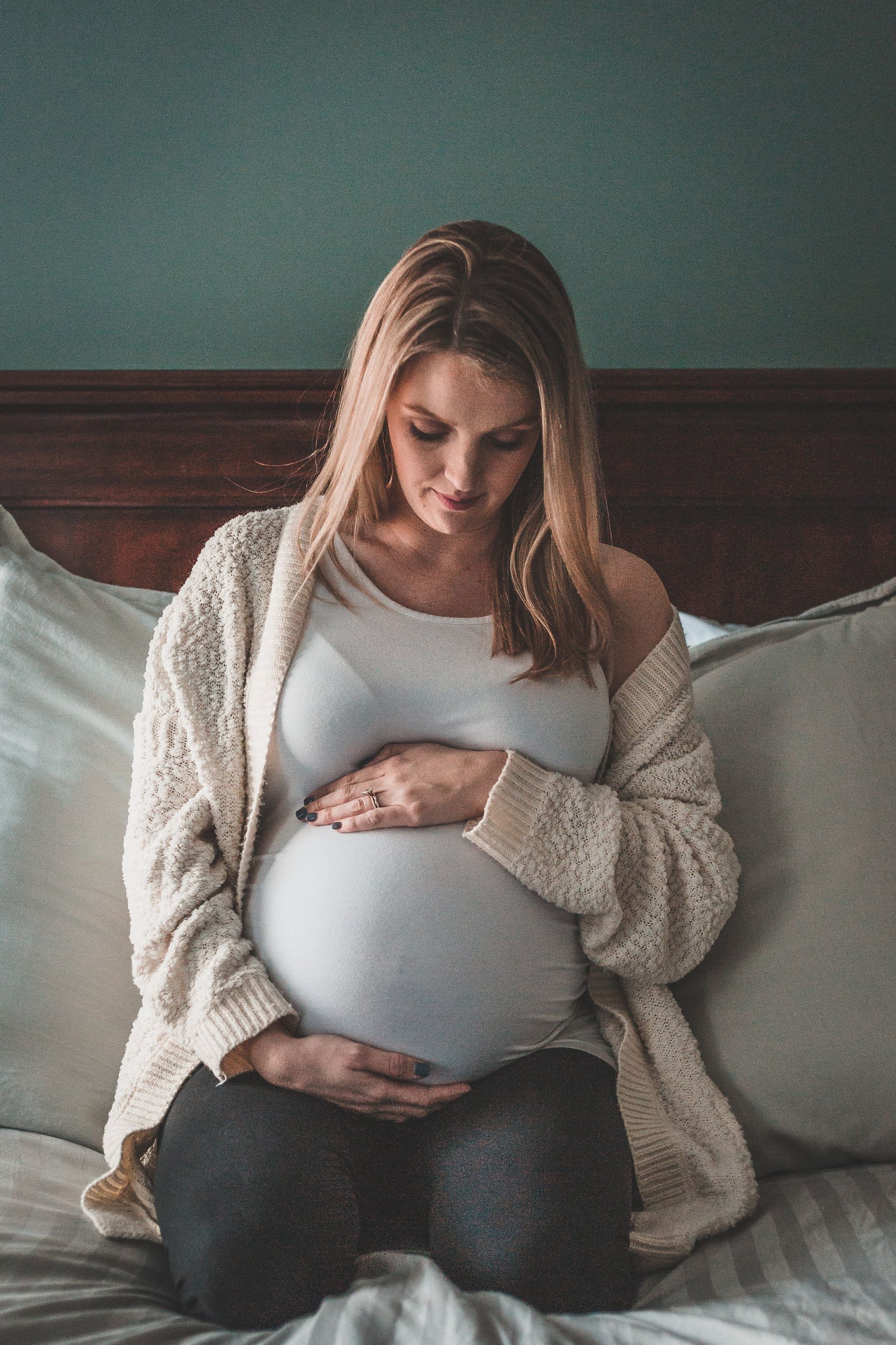 A pregnant woman is sitting on a bed holding her belly.