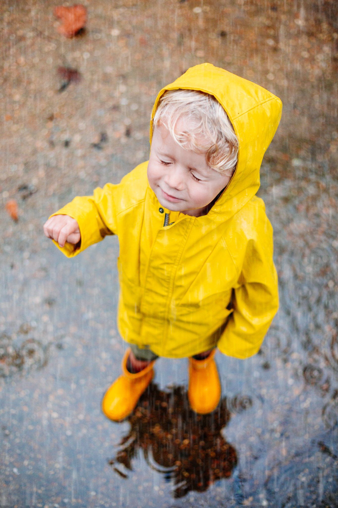 A little boy wearing a yellow raincoat and orange rain boots is standing in a puddle.