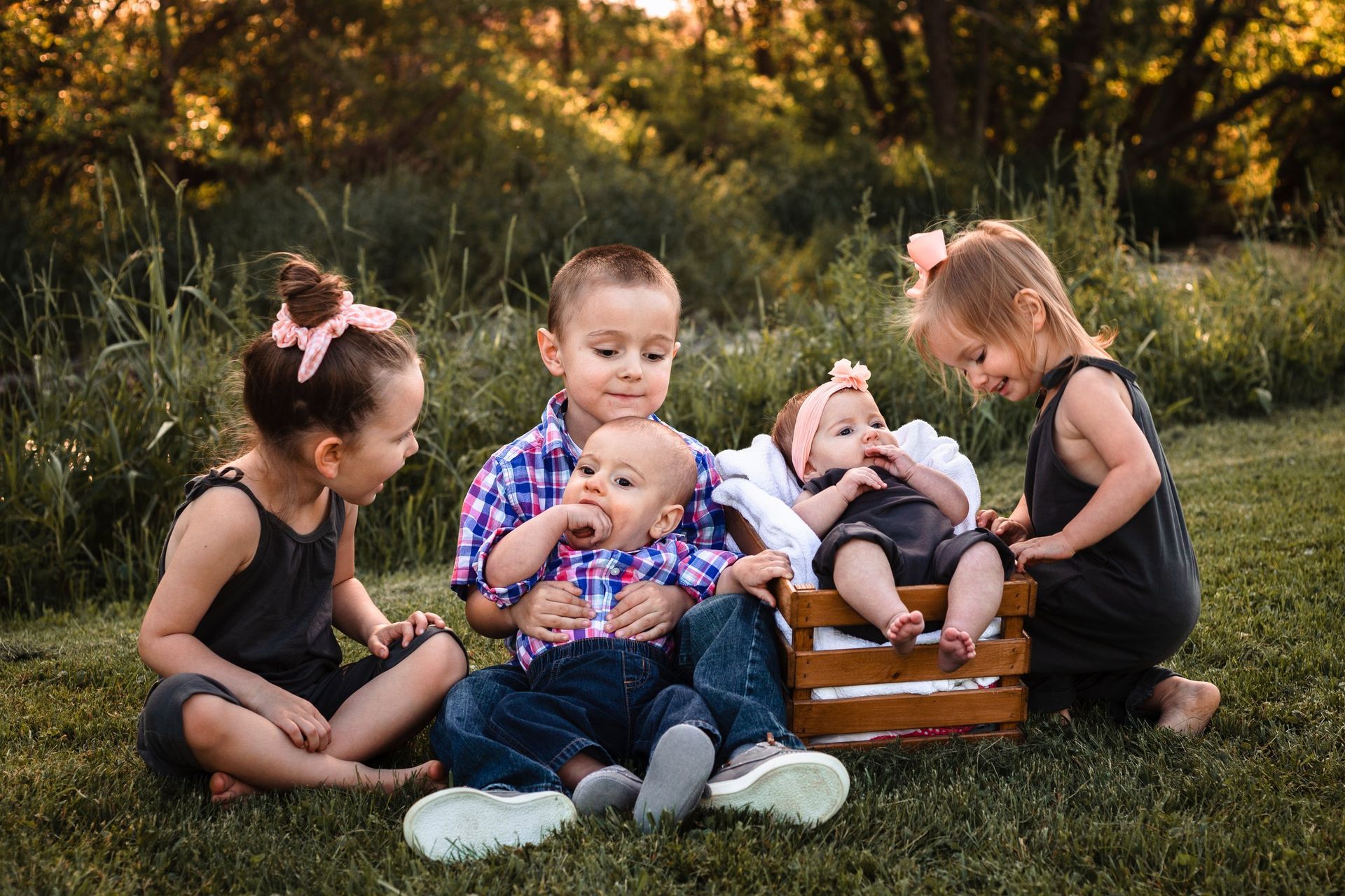 A group of children are sitting in the grass with a baby in a wooden crate.