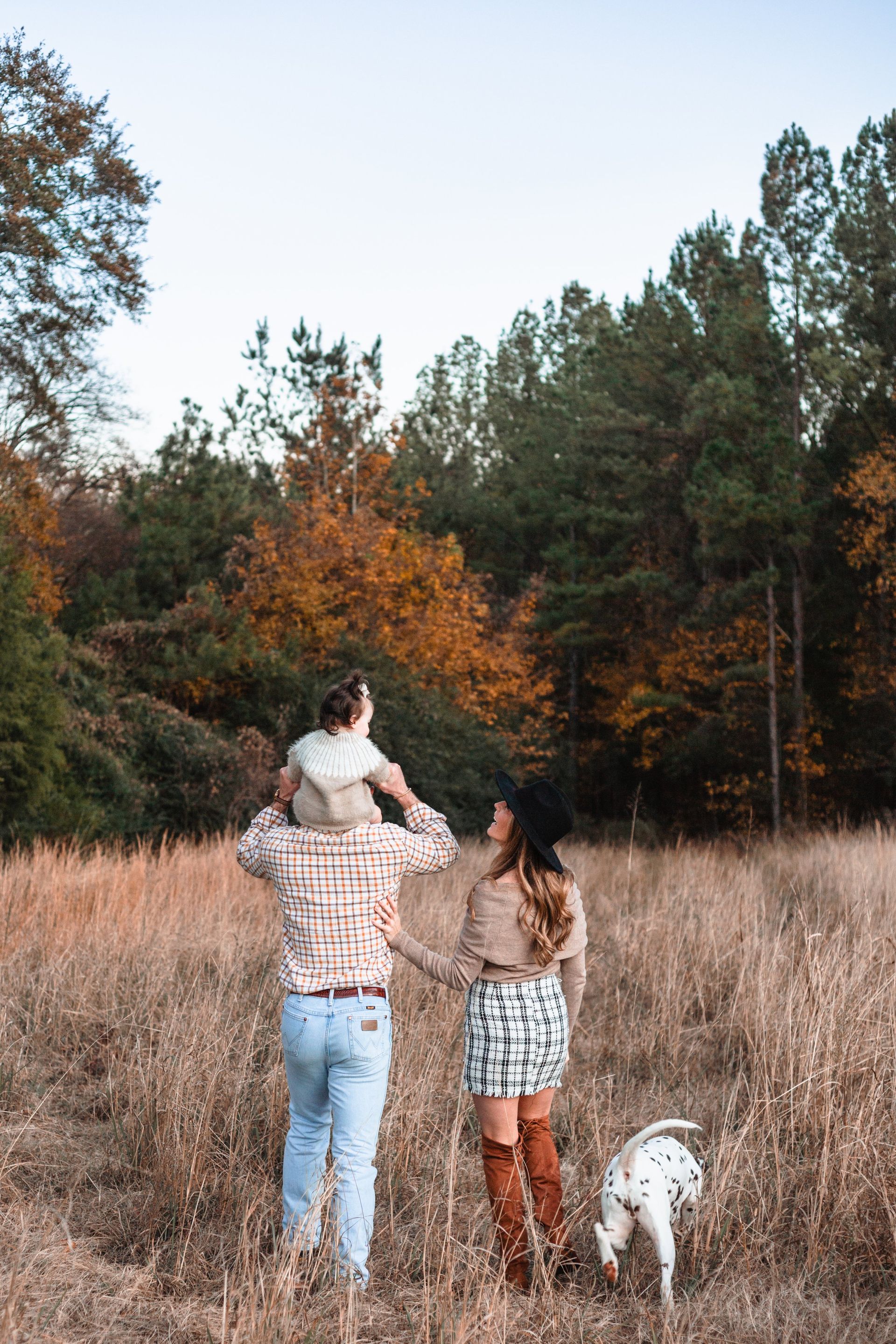 A family is standing in a field with a dog and a baby on their shoulders.