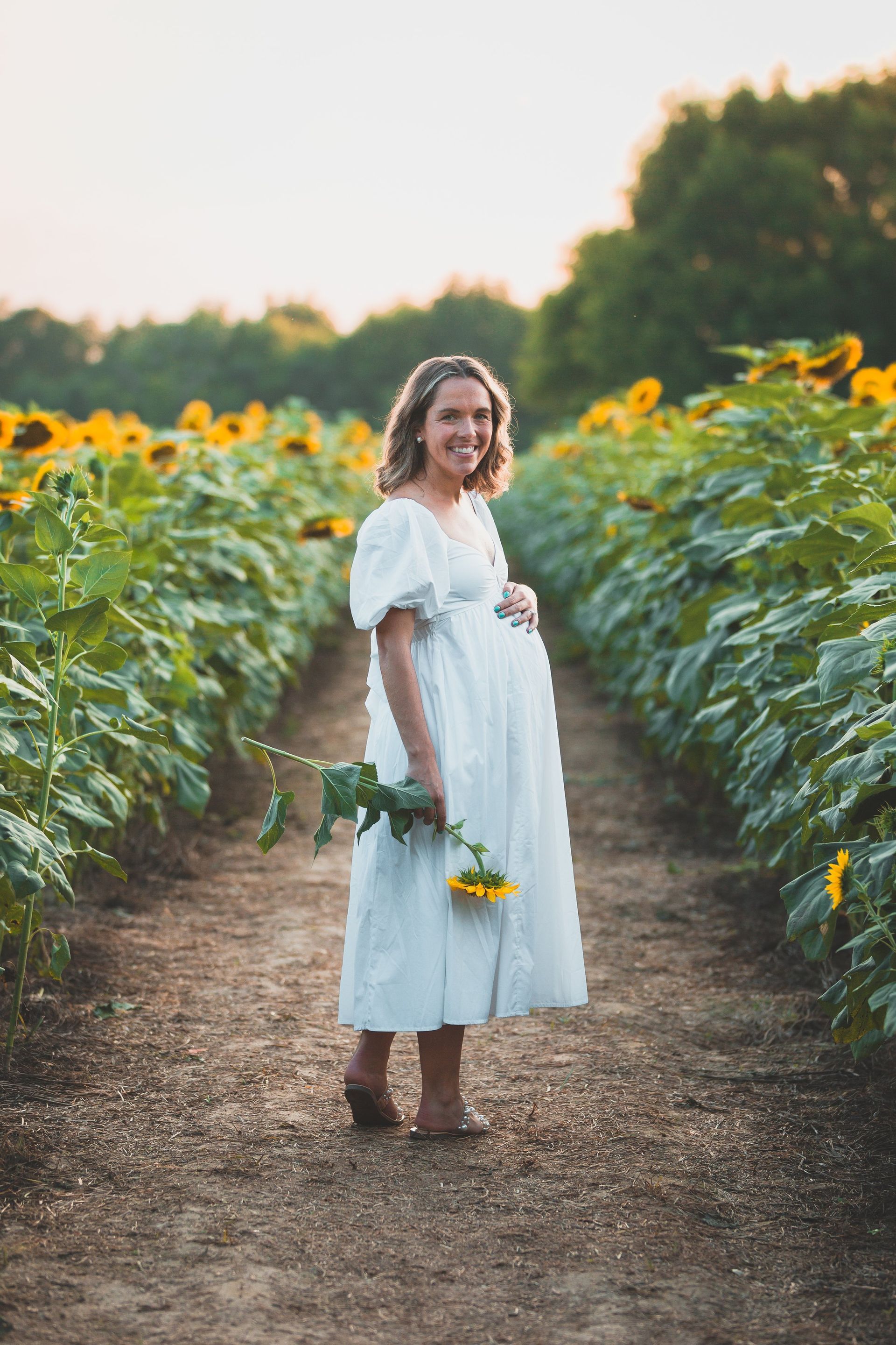 A pregnant woman in a white dress is standing in a field of sunflowers.