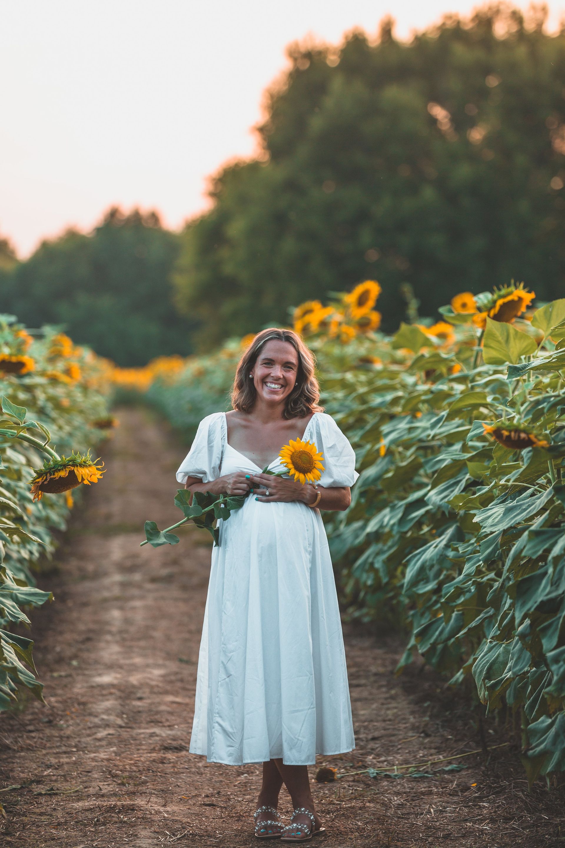 A woman in a white dress is standing in a field of sunflowers holding a sunflower.