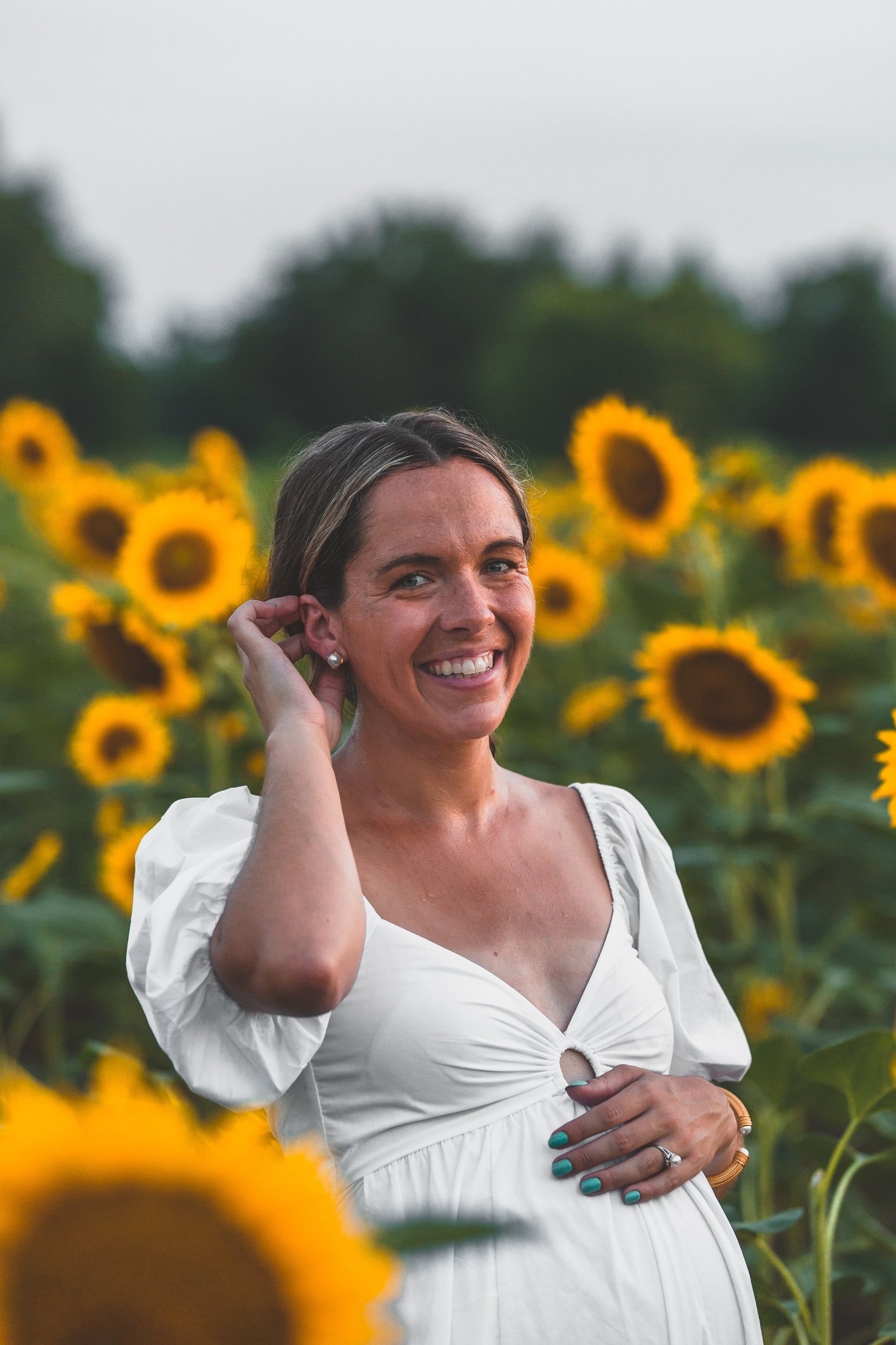 A pregnant woman in a white dress is standing in a field of sunflowers.