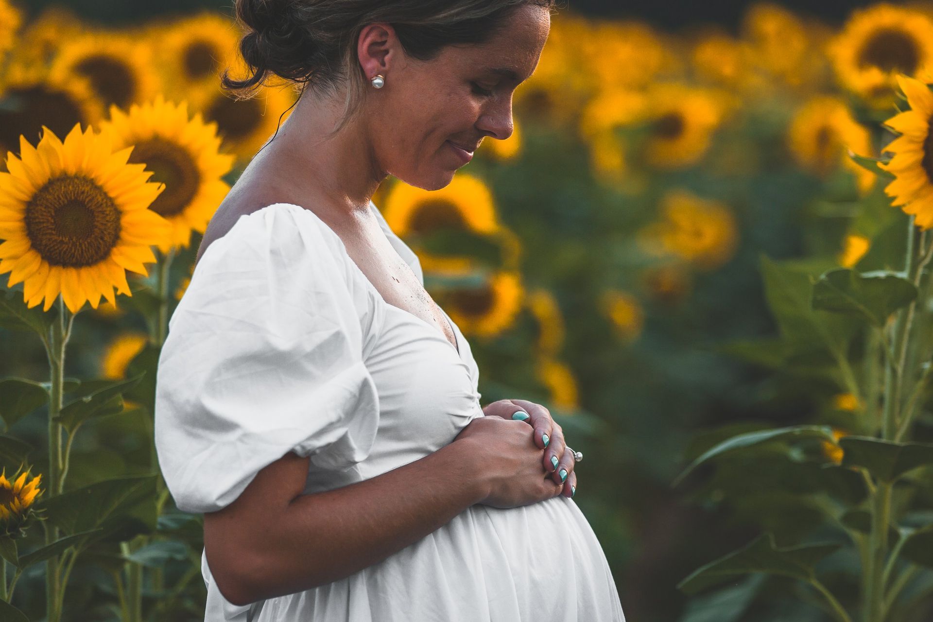 A pregnant woman in a white dress is standing in a field of sunflowers.