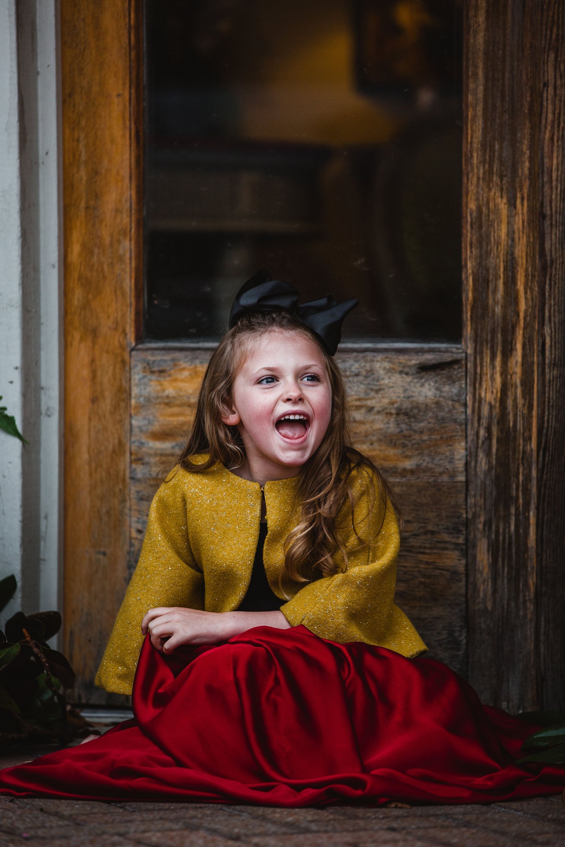 A little girl in a yellow jacket and red skirt is sitting on the ground in front of a door.