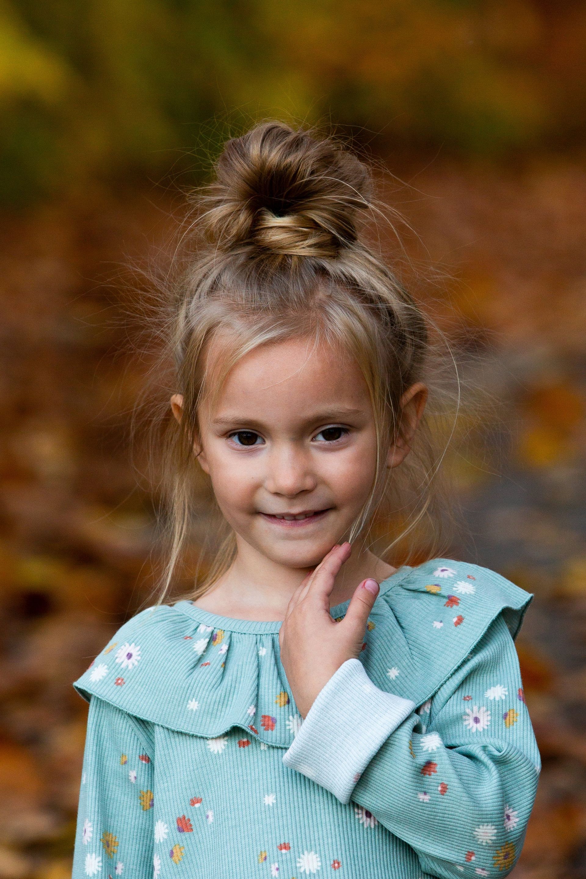 A little girl in a blue dress is standing in front of a pile of leaves.