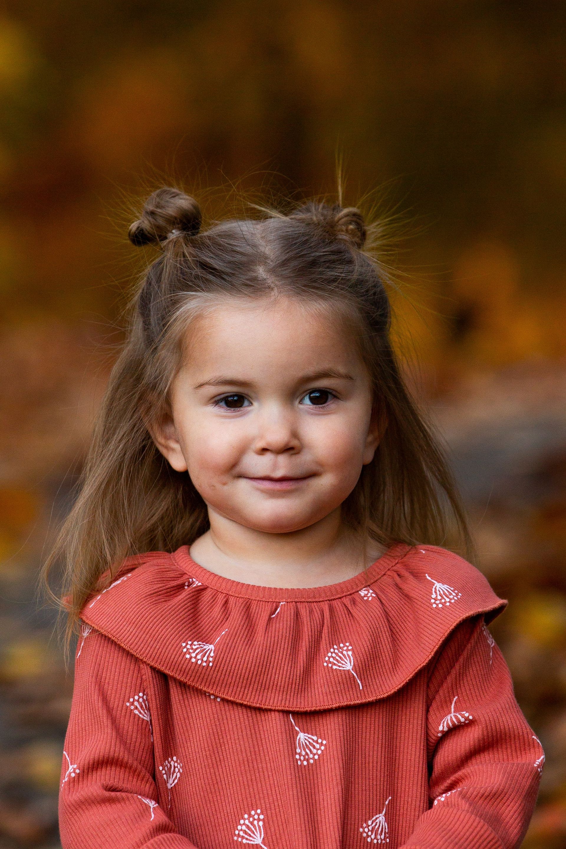 A little girl in a red dress is standing in front of a pile of leaves.