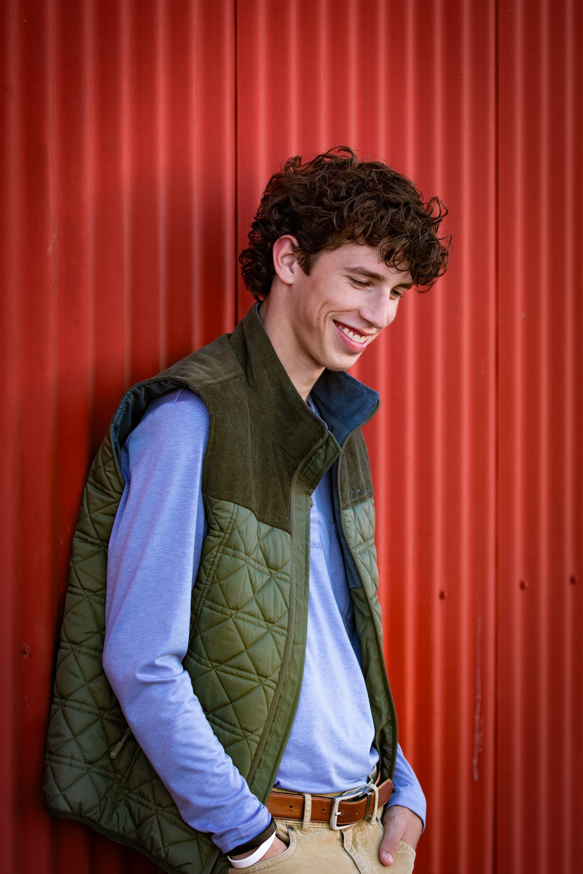A young man wearing a green vest and a blue shirt is standing in front of a red wall.