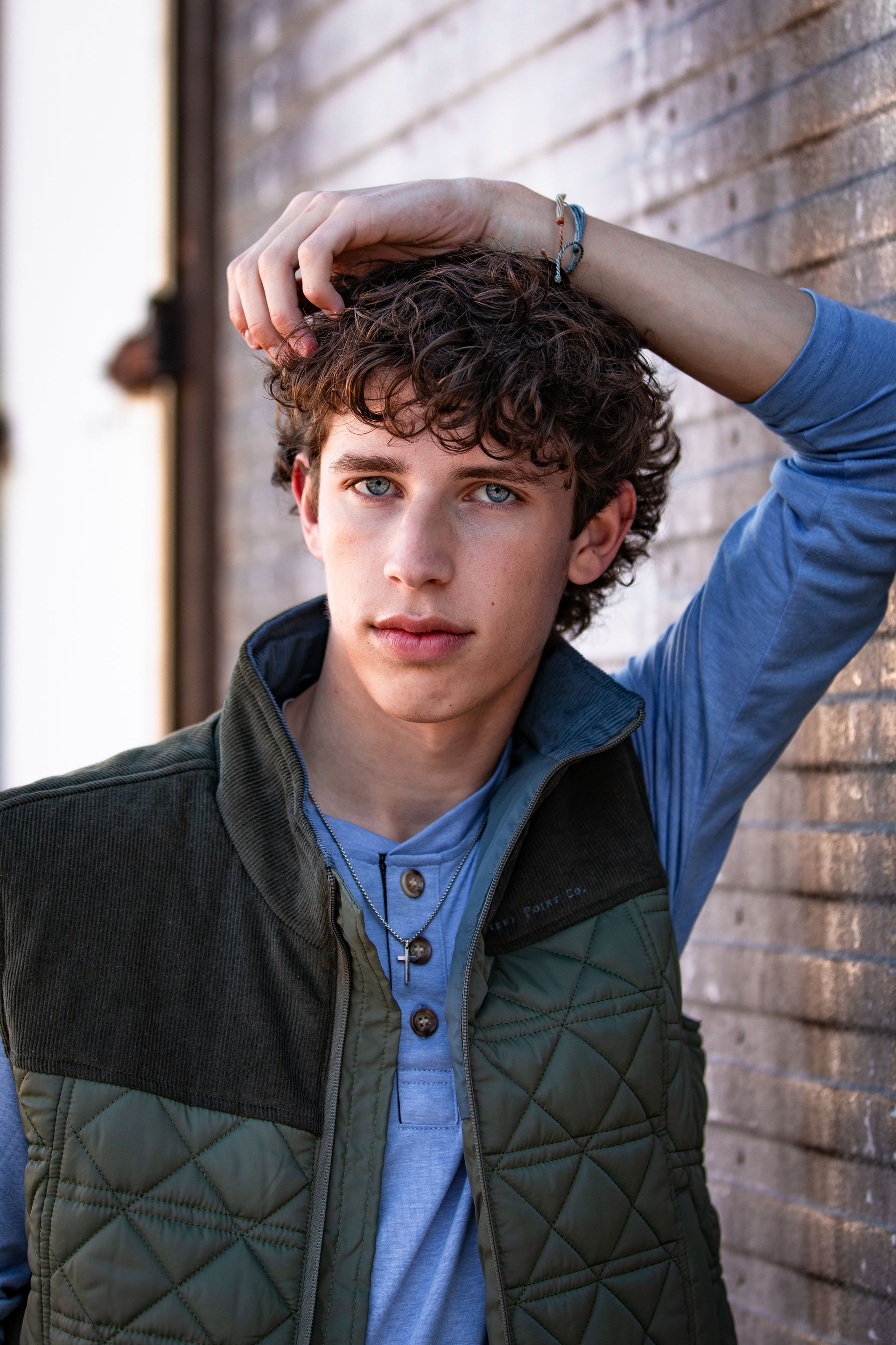 A young man wearing a vest and a blue shirt is standing in front of a brick wall.