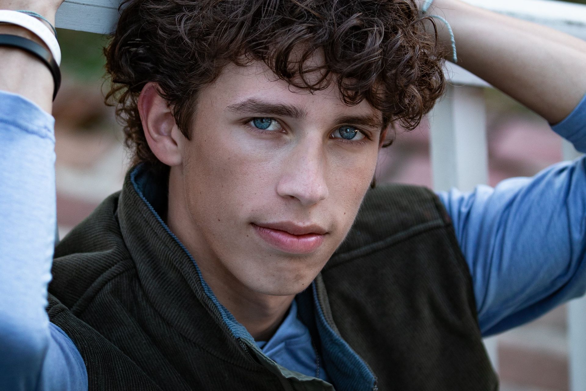 A young man with curly hair is wearing a vest and a blue shirt.