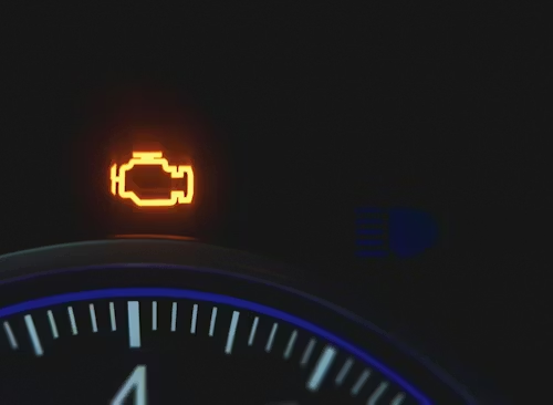 An illuminated orange check engine light on a car dashboard gauge cluster.