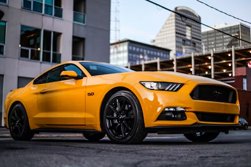 Yellow Ford Mustang parked on a city street, black wheels, modern buildings in the background.