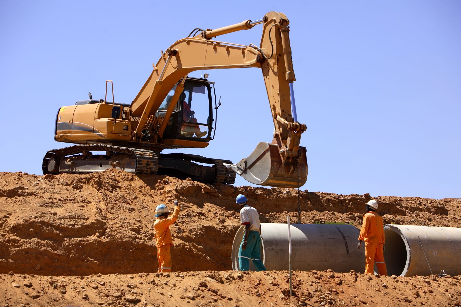 Un escavatore scava terra in un cantiere edile con operai in tute arancioni vicino a un grande tubo