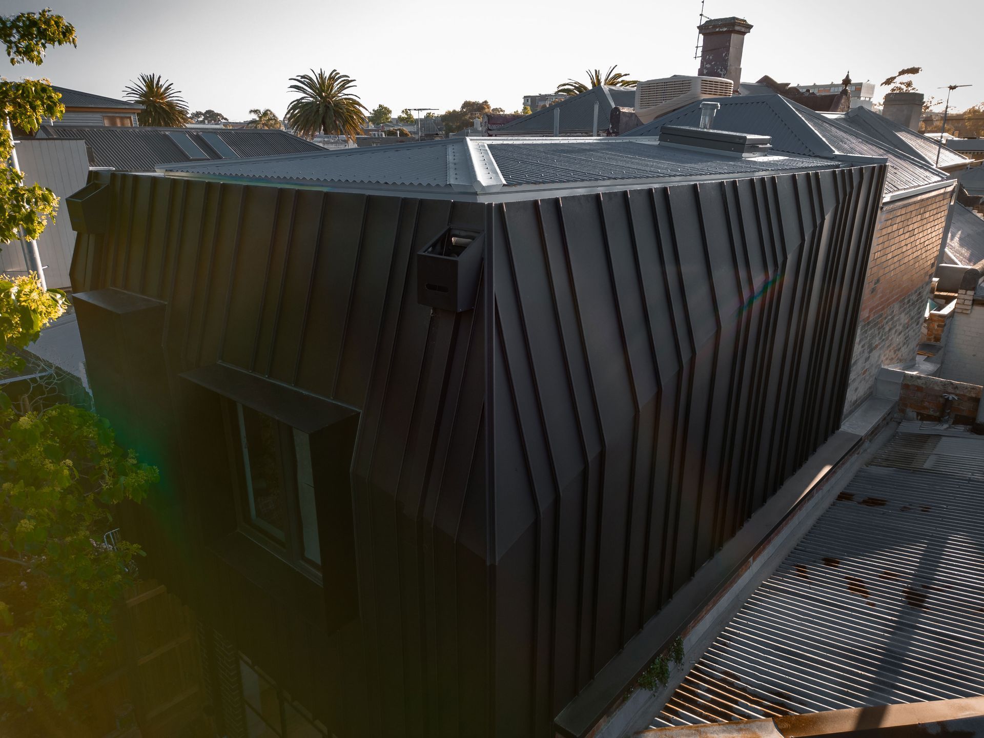 Dark metal roof and walls of a modern building, viewed from above, with other roofs and trees in the background.