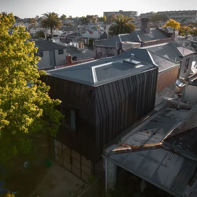 Black metal-clad modern house with rooftop views, surrounded by traditional homes and trees.
