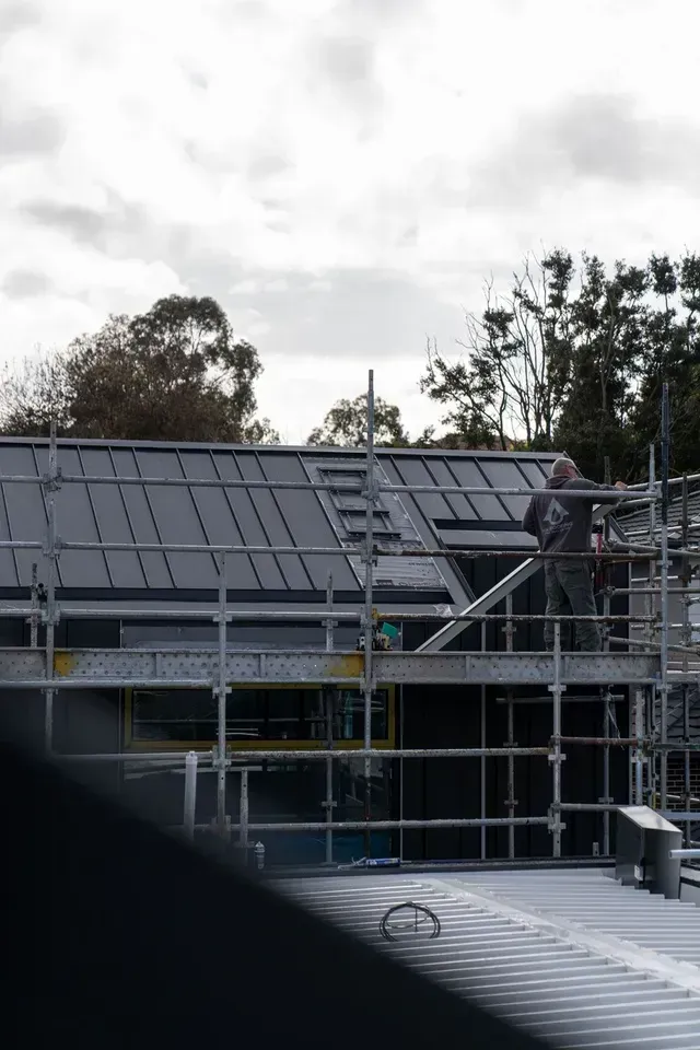 A construction worker on scaffolding near a building roof. He's wearing a black uniform, working with tools.