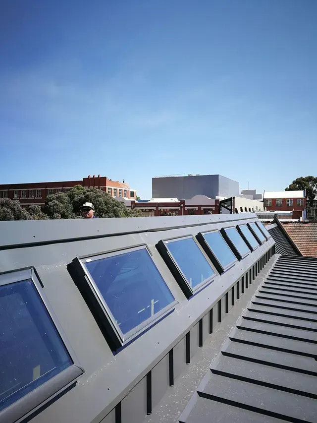 Row of skylights on a dark gray roof against a clear blue sky. Buildings and trees in the background.