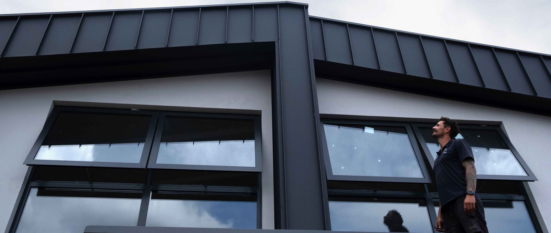 Man looking upwards near open windows of a modern building with a dark roof and cloudy sky.