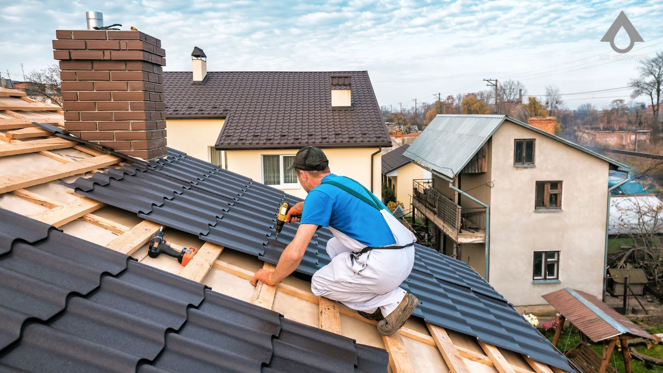 Roofer on a roof installing dark metal tiles with a power drill; houses in the background.