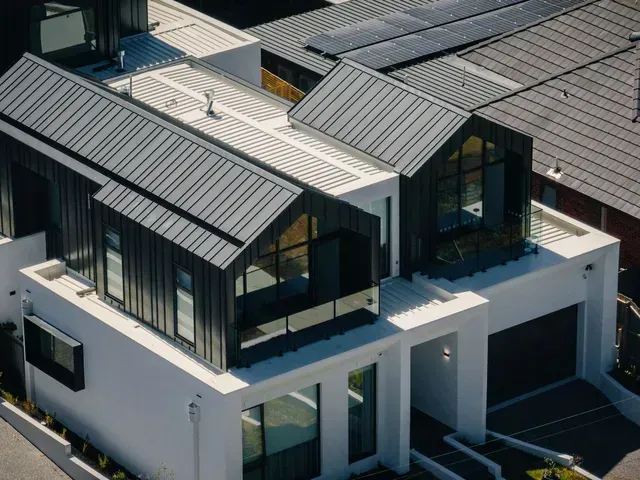 Modern two-story house with dark metal roof, black-framed windows, white exterior, and solar panels.