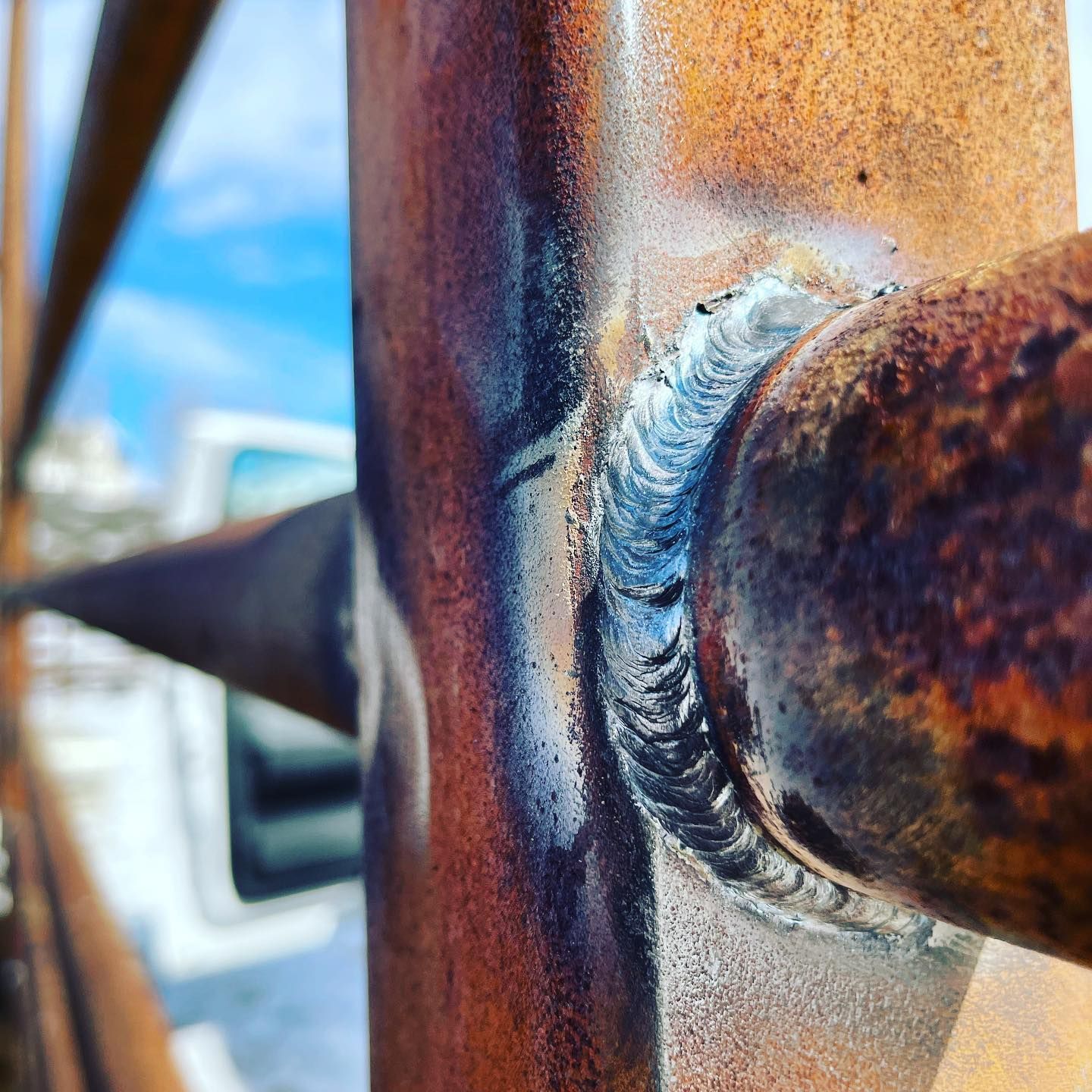 A close-up view of a clean, silver-colored metal weld joining a rusted round pipe to a square post outdoors.