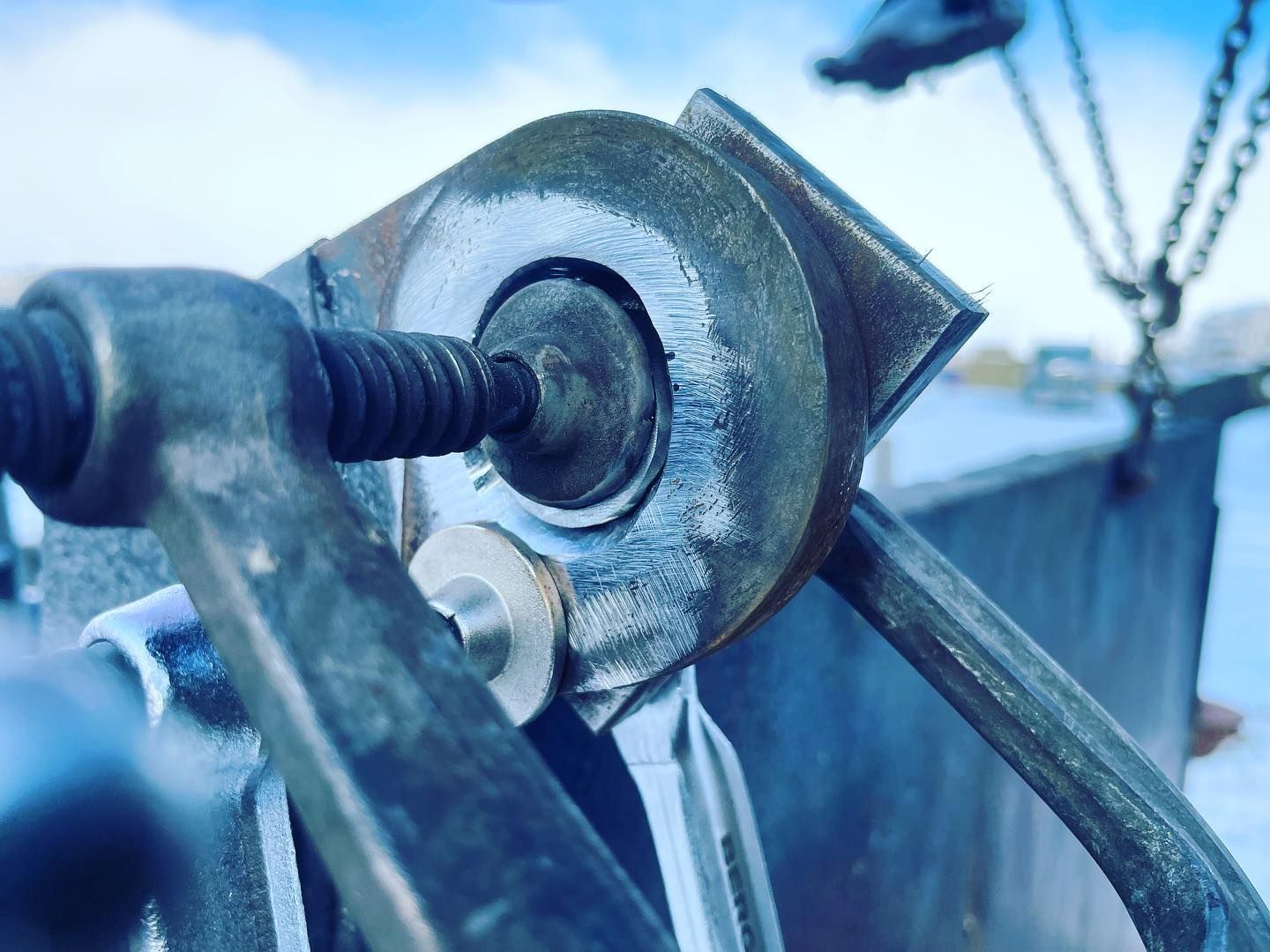 A close-up of a metal clamp tightening a bolt on a circular industrial plate, with a chain hoist visible in the background.