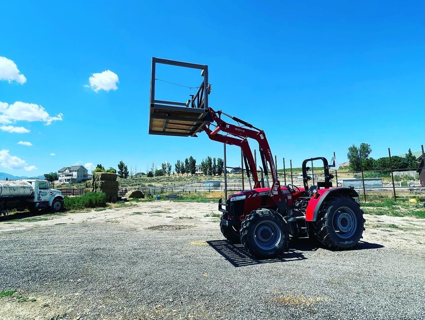 A red tractor equipped with a raised metal work platform sits on a gravel lot under a clear blue sky.