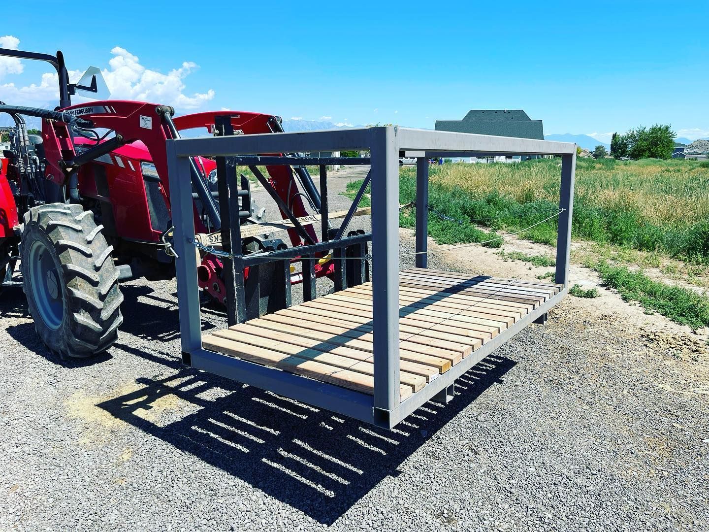 A red tractor with a front-end loader attached to a gray, rectangular metal cage with a wooden slatted floor.