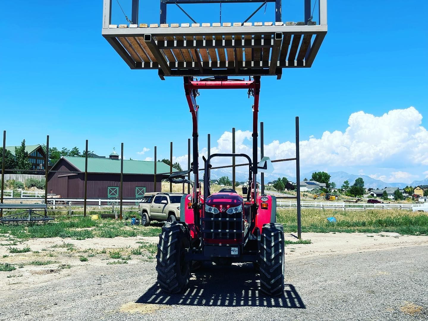 A red tractor with a large elevated wooden platform attachment sits on a gravel lot under a clear blue sky.