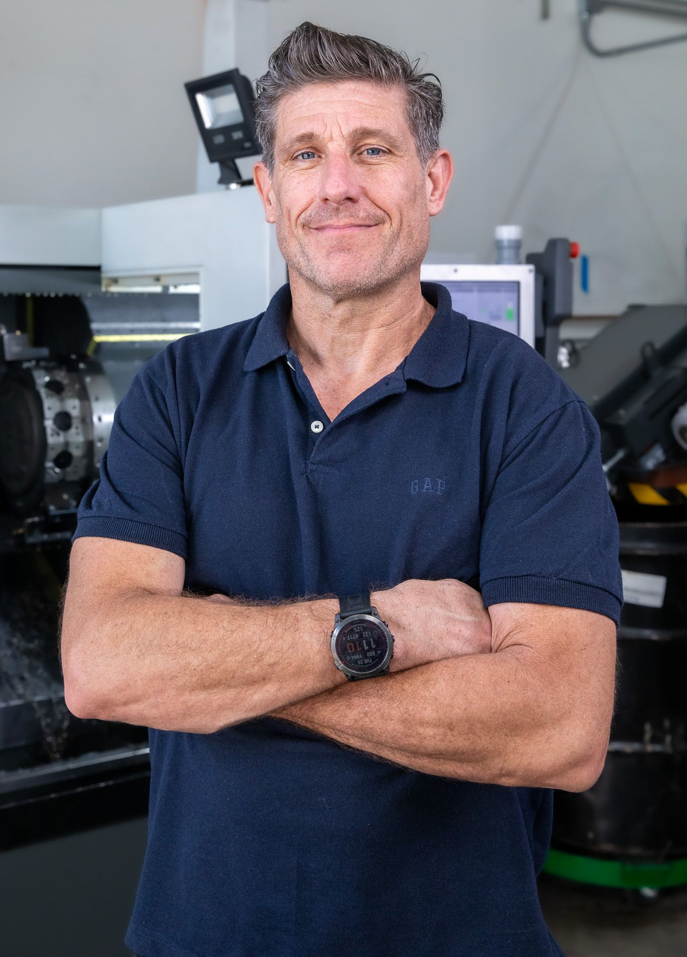 John Carter of Precision Metal Craft with arms crossed, standing in a workshop, smiles.