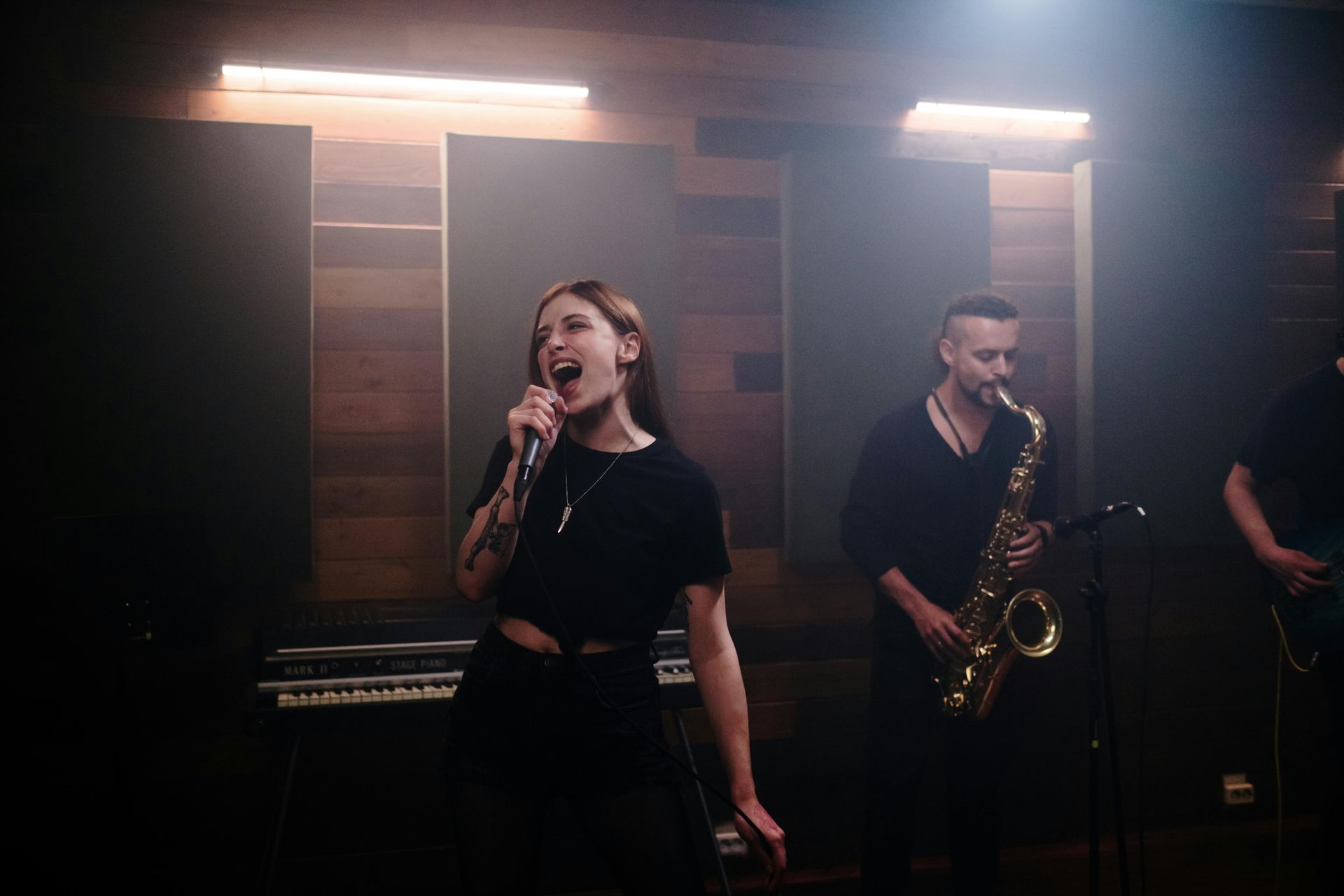 A singer performing into a microphone alongside a saxophonist in a dimly lit studio with wood-paneled walls.