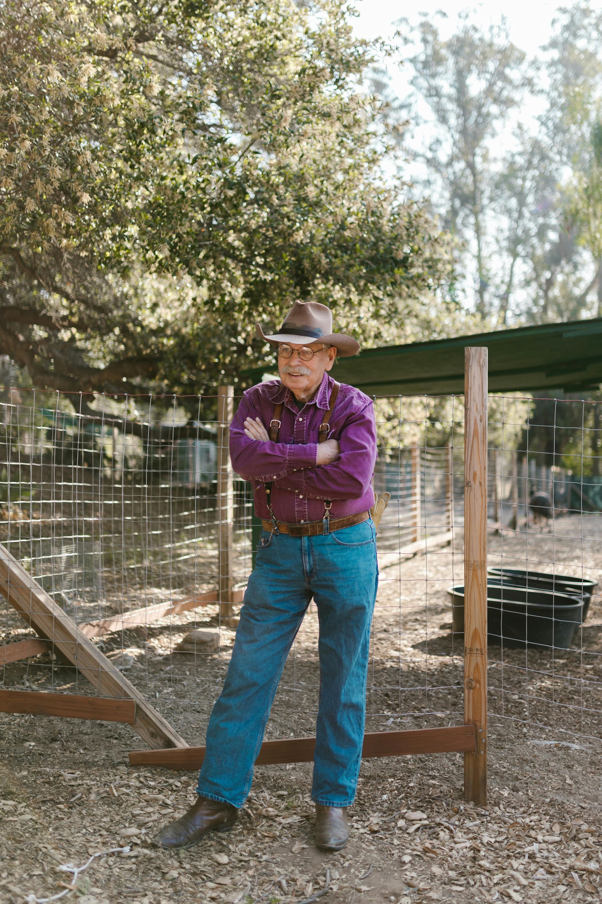 A person wearing a purple shirt, blue jeans, and a cowboy hat stands with arms crossed in a fenced, wooded outdoor area.