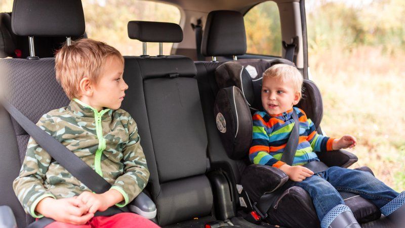 Two children sit in the back seat of a car, one in a camouflage jacket and the other in a striped shirt and car seat.