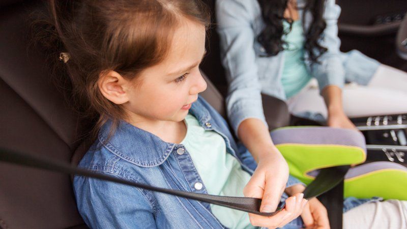 A person helps a child fasten a seatbelt while sitting in a car with a lime green and grey booster seat.