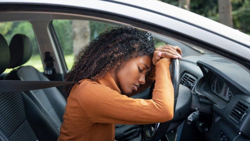 A woman is sleeping in a car with her head on the steering wheel.