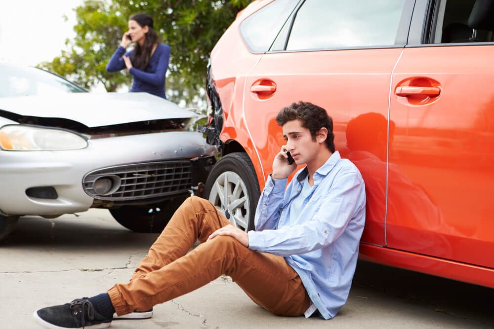 A man is sitting on the ground talking on a cell phone in front of a car accident.