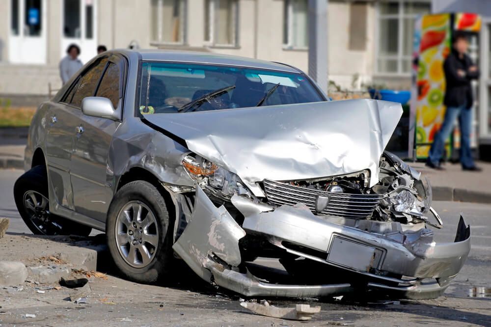 A silver sedan sits on a roadside with extensive front-end damage and a buckled hood, likely from a collision.