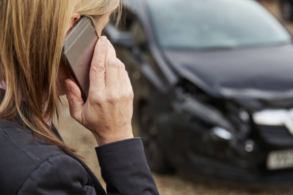 A person speaking on a smartphone in front of a dark car with front-end damage.