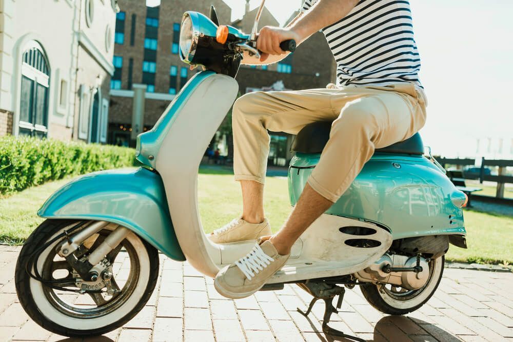 A man is riding a blue and white scooter on a brick sidewalk.