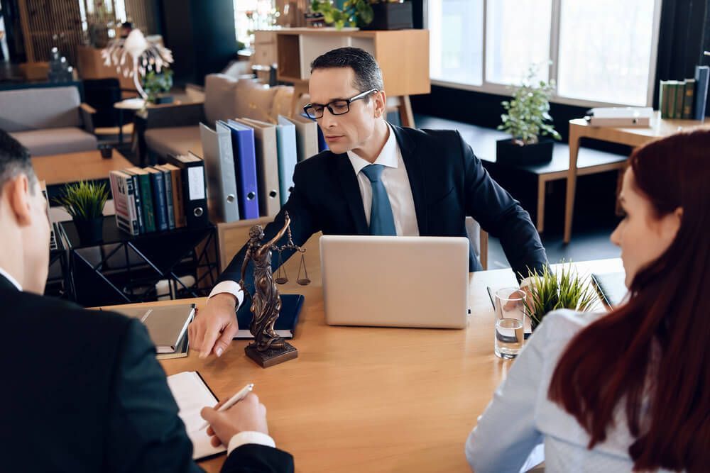 A man in a suit and tie is sitting at a desk talking to two people.