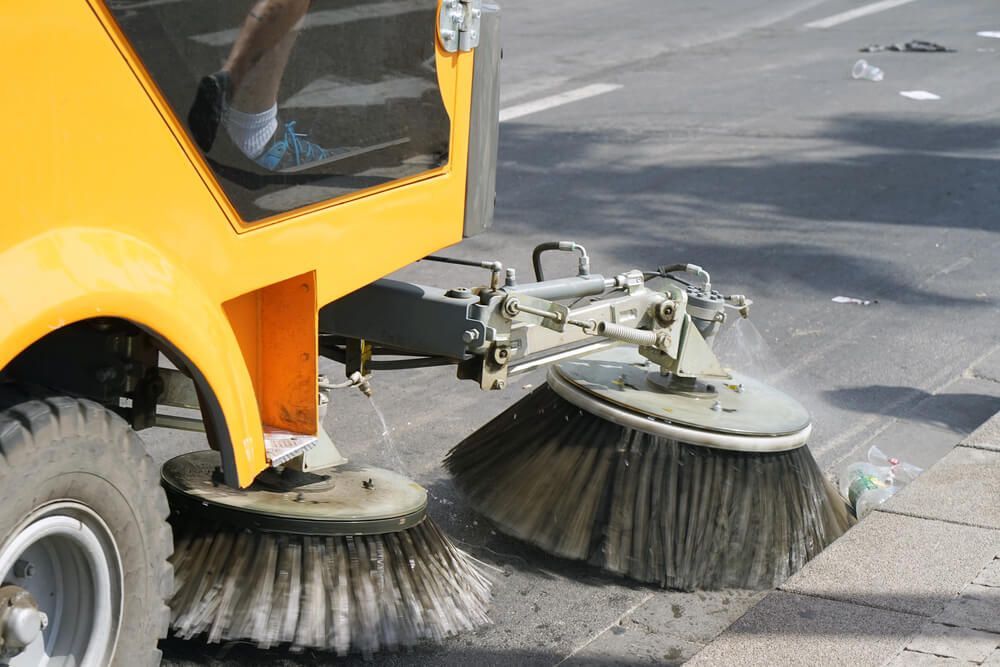 A yellow street sweeper is cleaning the street with brushes